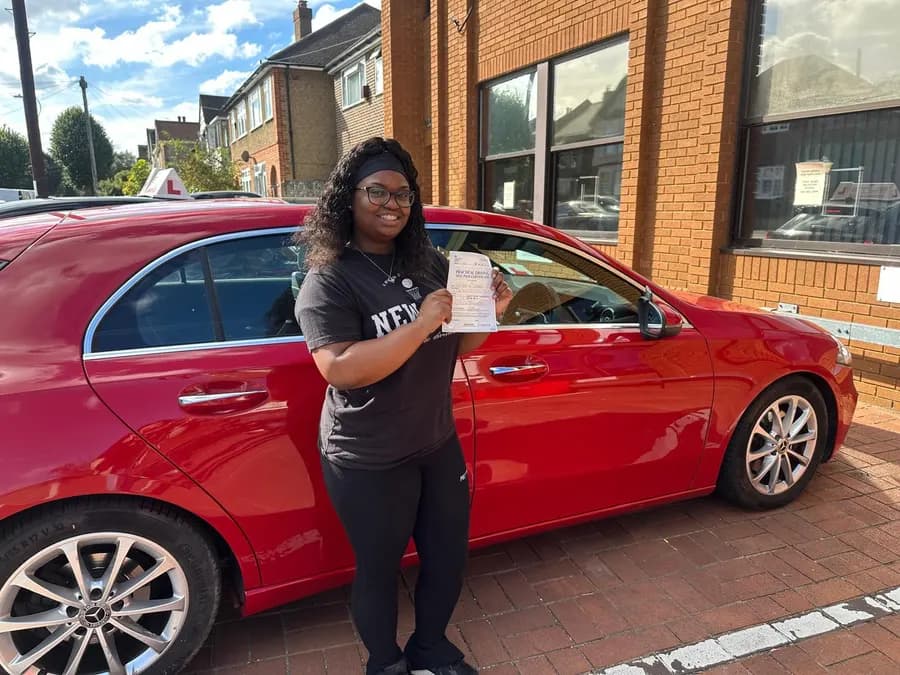 Smiling learner driver holding her driving test pass certificate beside a red Mercedes A-Class tuition car