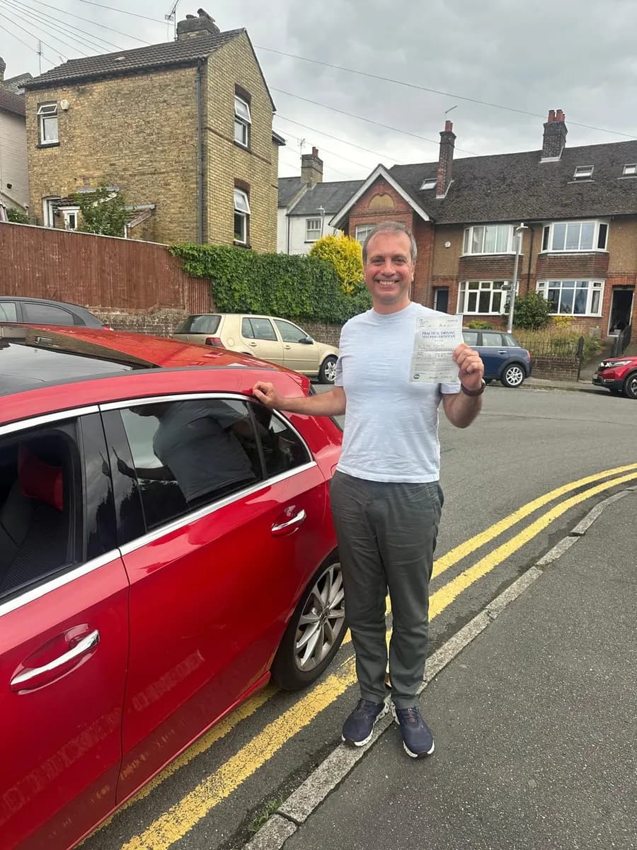 Learner driver holding his pass certificate next to a red Mercedes tuition car on a quiet residential street