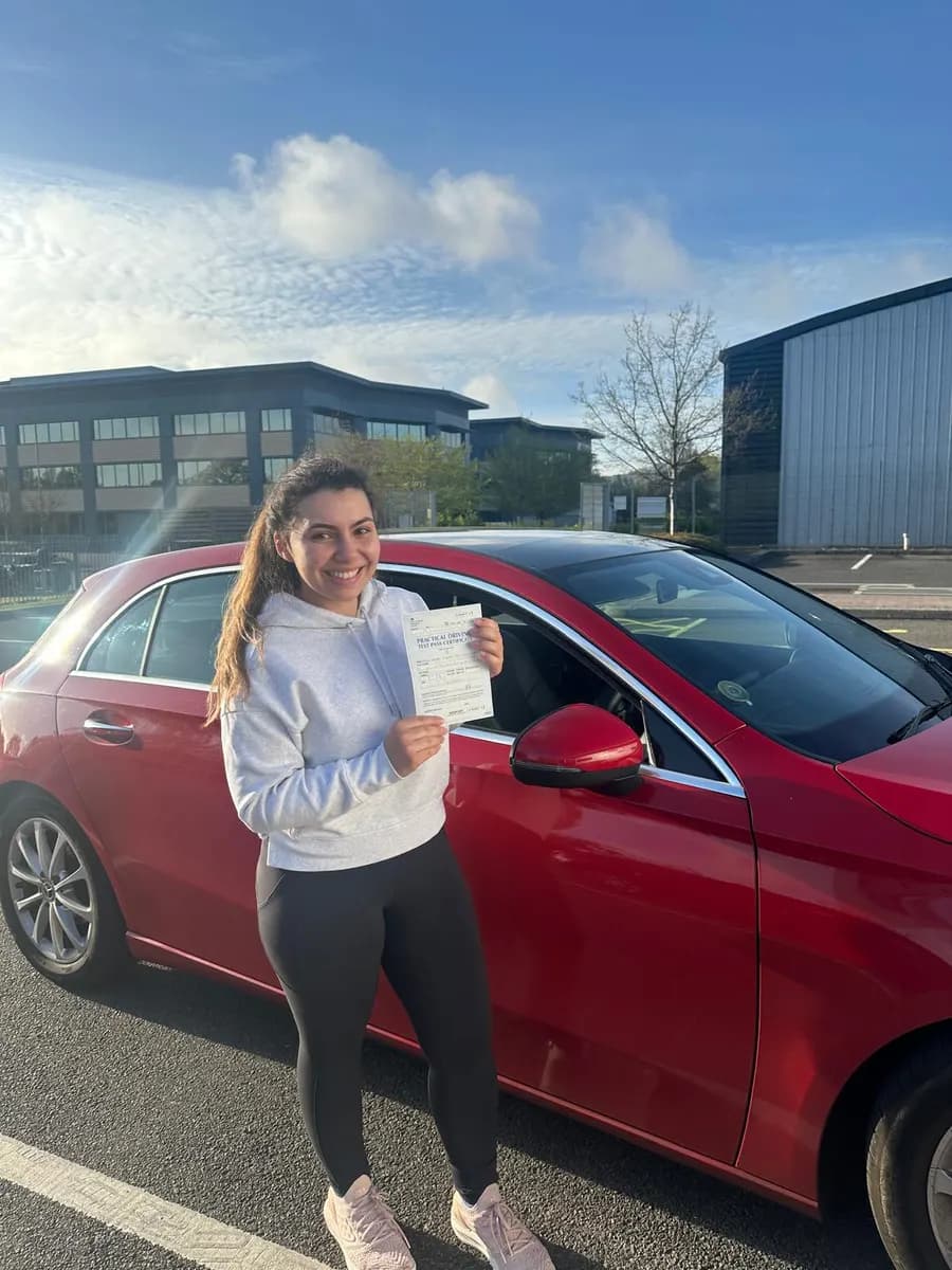 Happy student in a white hoodie showing her driving test pass certificate beside a red Mercedes A-Class