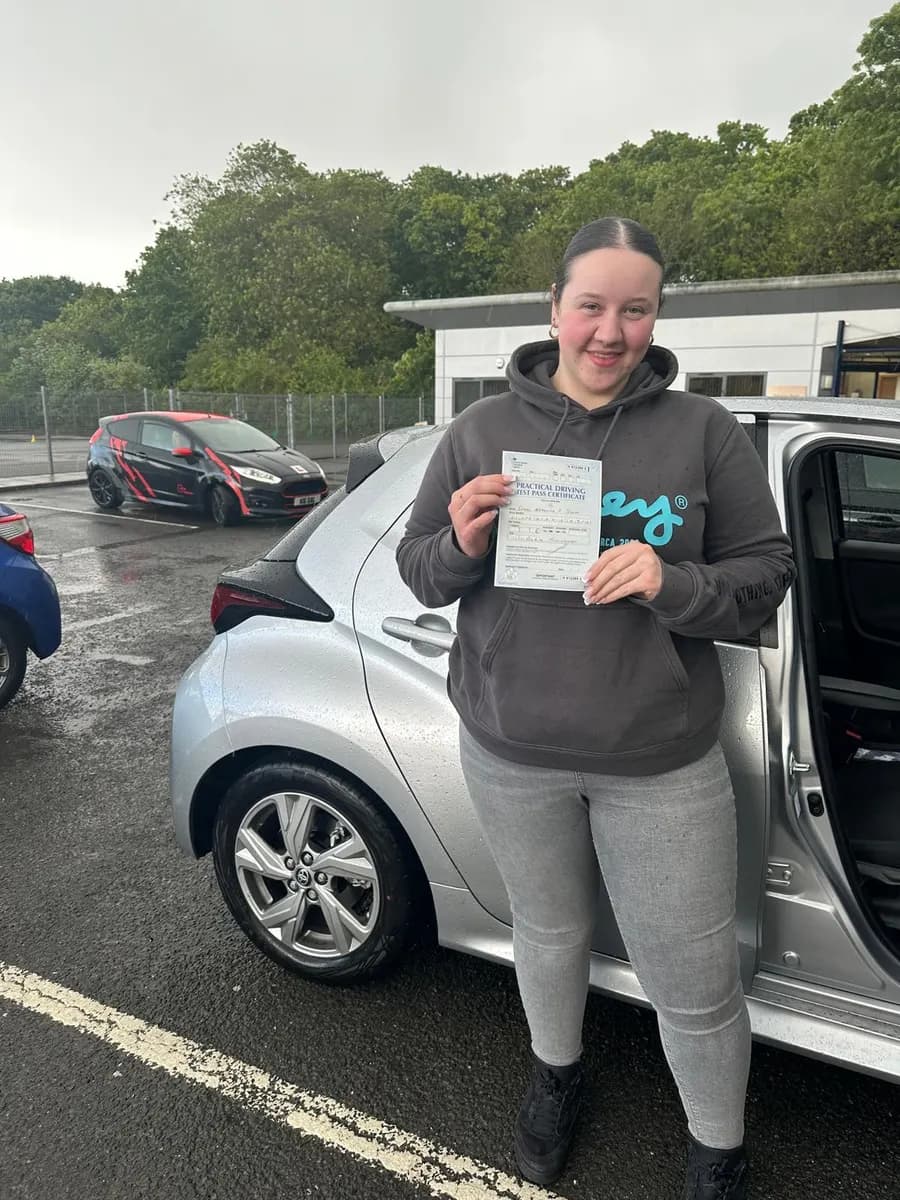 Student in a grey hoodie holding her driving test pass certificate next to a silver hatchback tuition car