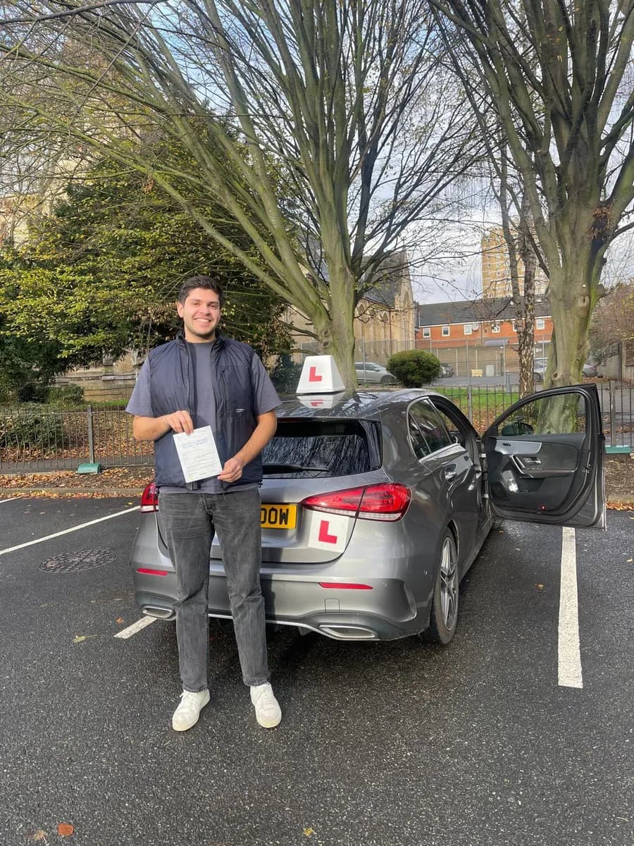 Young man holding his pass certificate behind a grey Mercedes driving school car with an L-plate roof sign