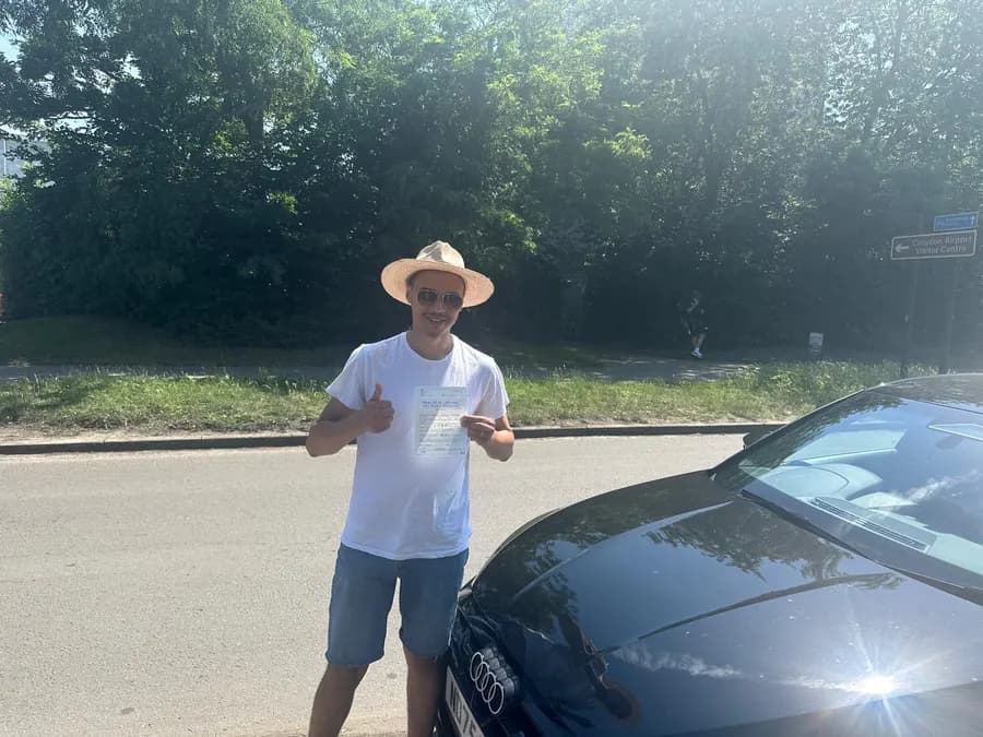 Man in a sun hat giving a thumbs up with his driving test pass certificate beside the car
