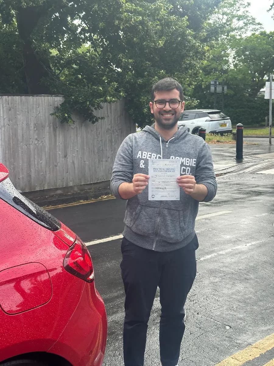 Learner in a grey hoodie holding his driving test pass certificate beside a red tuition car after a wet-weather test