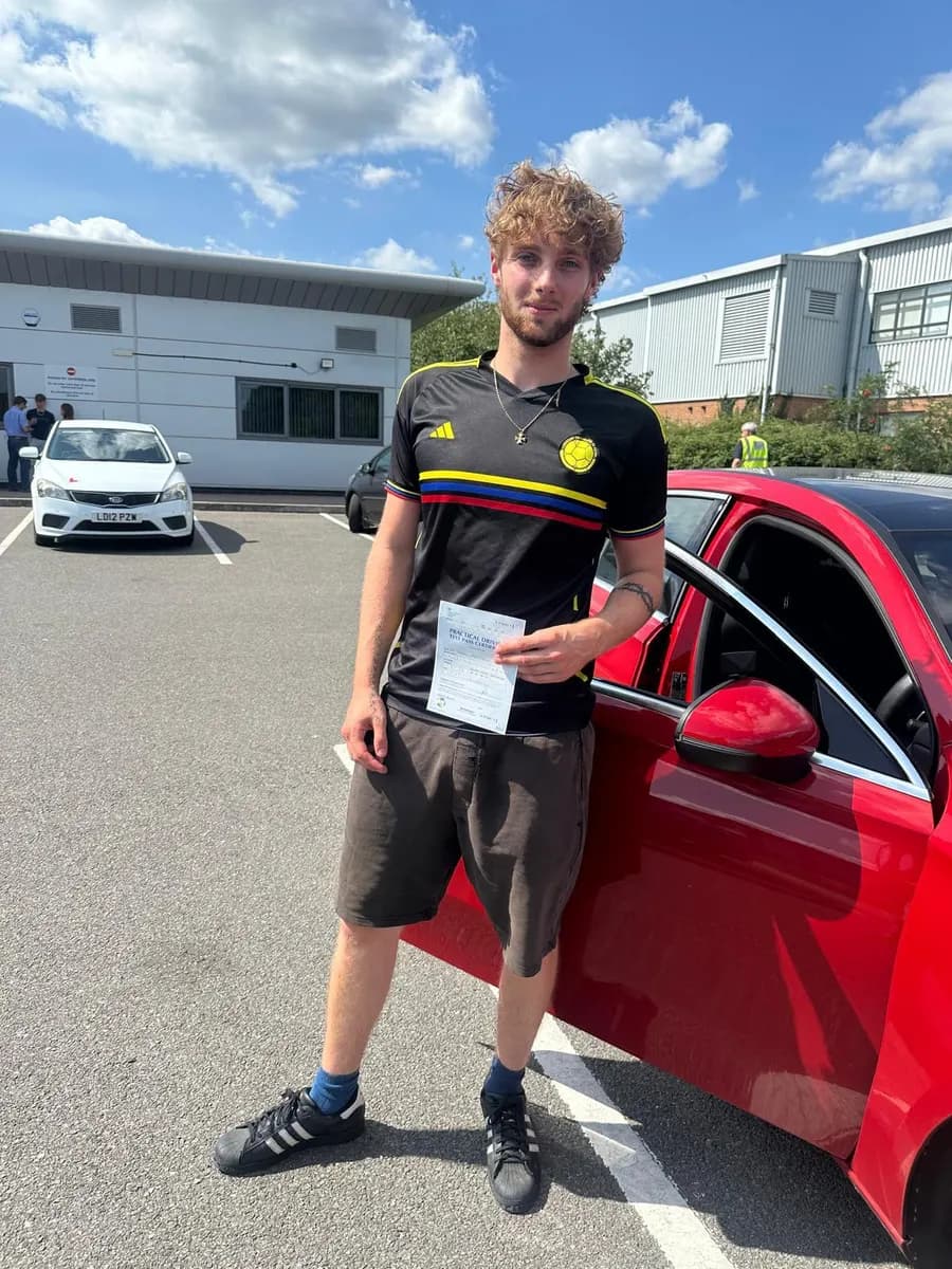 Young man holding his driving test pass certificate by the open door of a red tuition car